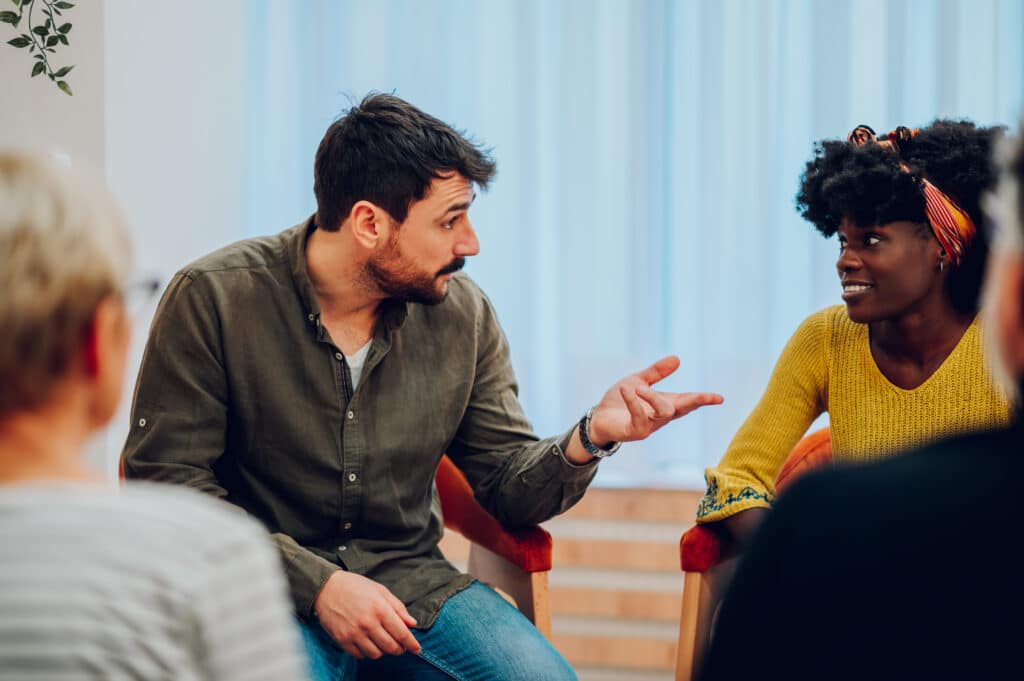 Man supporting african american woman new attender of group therapy to talk about herself during a meeting at mental health center. Group therapy participants sitting in a circle and talking.